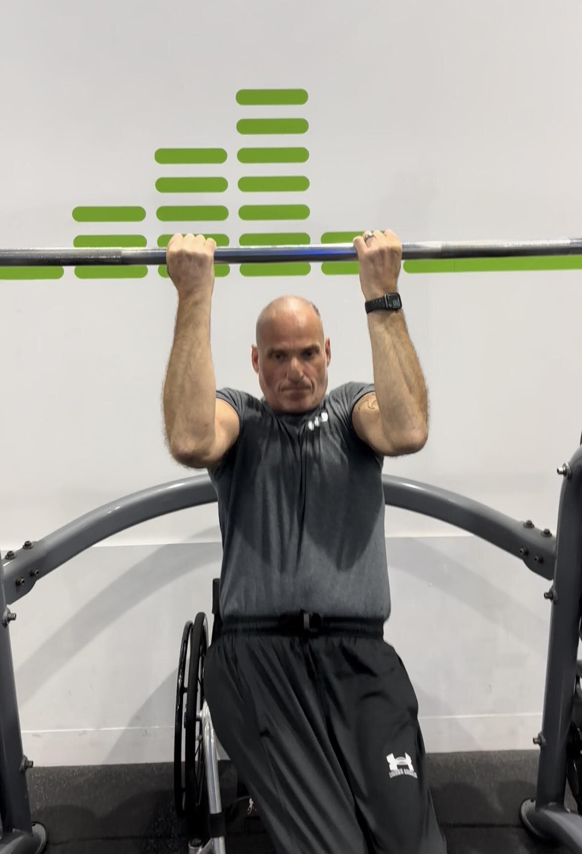 Matt Anderson in his wheelchair performing an overhead barbell press in a gym